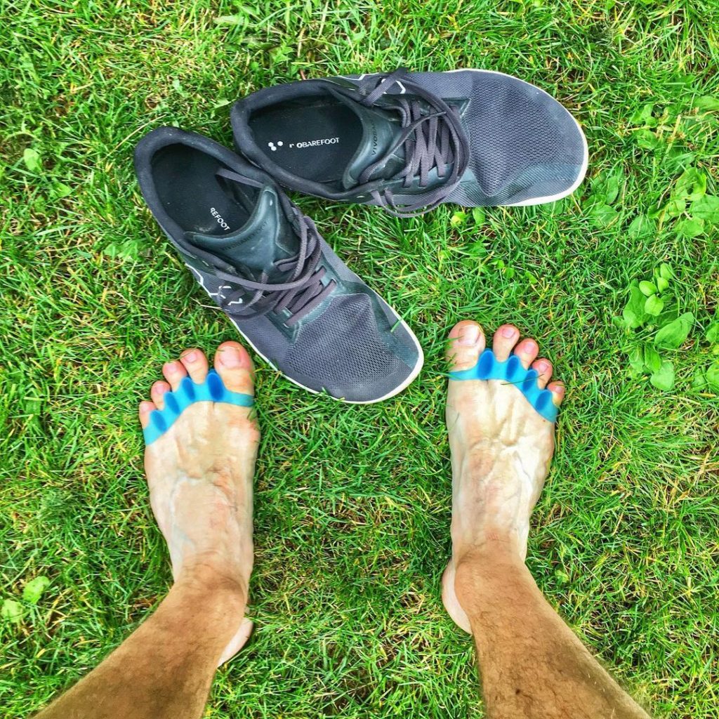 Men's feet with toe spacers next to barefoot shoes in grass.